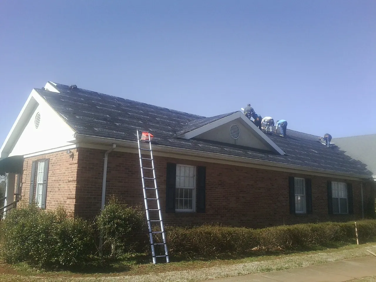 Crew tearing shingles off a brick ranch roof with a ladder set — Emergency Roof Repair work by our crew in O Fallon